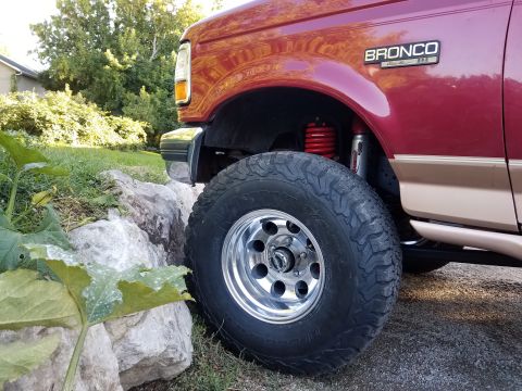 1995 Ford Bronco - The front quarter panel and wheel of a red and tan Ford Bronco are shown, with a prominent 'BRONCO Eddie Bauer' badge.