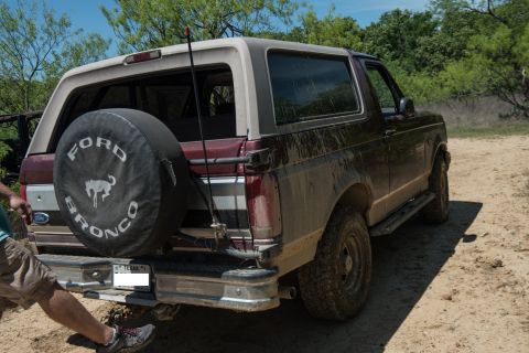 1996 Ford Bronco - Rear view of a muddy Ford Bronco parked on a dirt road with trees in the background.
