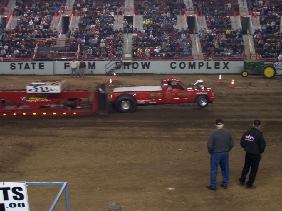 Red dually pickup truck pulling a weighted sled in a dirt arena at a State Farm Show Complex event