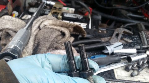1981 Ford Bronco - A close-up view of two bolts being held, with various tools and engine parts visible in the blurred background.