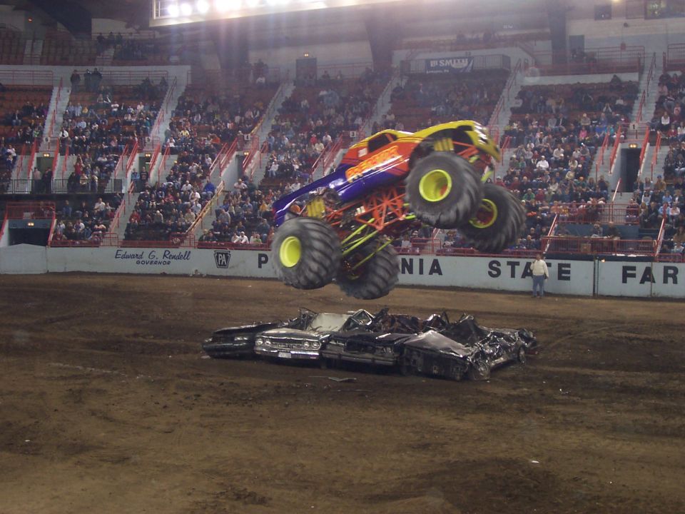 Orange and purple monster truck airborne above a crushed car in a dirt arena