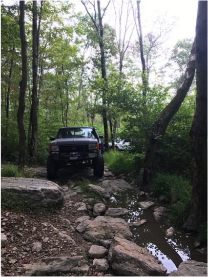 1989 Ford Bronco - A dark-colored Ford Bronco navigates a rocky, wooded trail with a stream running alongside it.