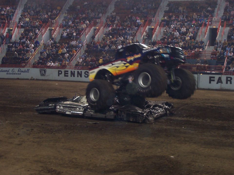 Orange and blue monster truck crushing a sedan flat in a dimly lit dirt arena