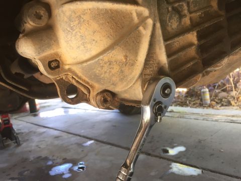 1986 Ford Bronco - Working on a transmission housing with a ratchet wrench, fluid visible on the garage floor.
