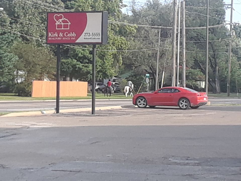 Red sports car parked on street with a real estate sign and horses in background