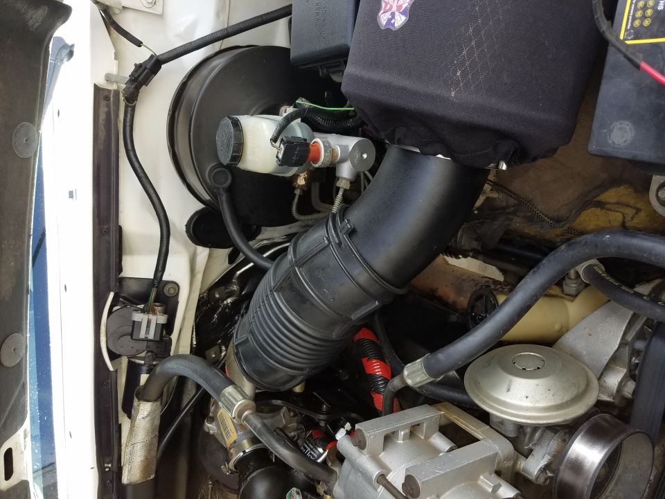 Engine bay of white pickup showing black intake tube, brake master cylinder, booster, and covered air filter