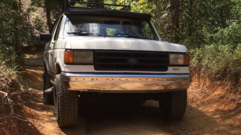 1988 Ford Bronco - This white Ford Bronco is driving on a dirt path surrounded by trees.