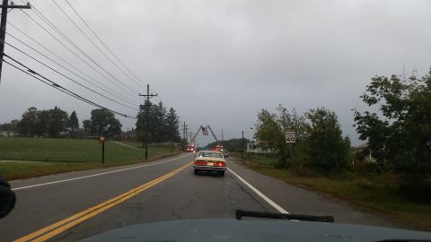 1991 Ford Crown Victoria - A car drives down a wet road under a cloudy sky, passing a fire truck displaying an American flag.