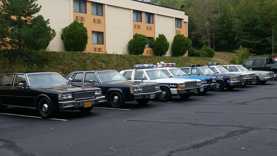 Row of police cars parked in a lot, including a black sedan and several marked units with light bars