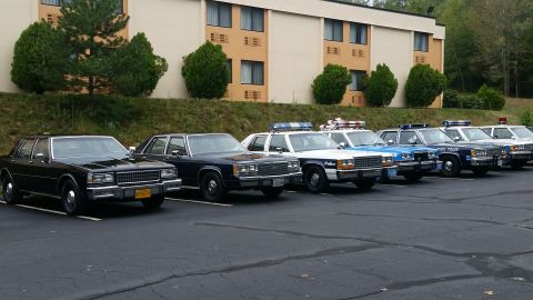 1991 Ford Crown Victoria - Several police vehicles are parked in a lot in front of a motel, including a black sedan and multiple marked patrol cars.