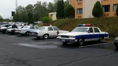 1991 Ford Crown Victoria - Multiple police vehicles are parked in a lot next to a building and trees.