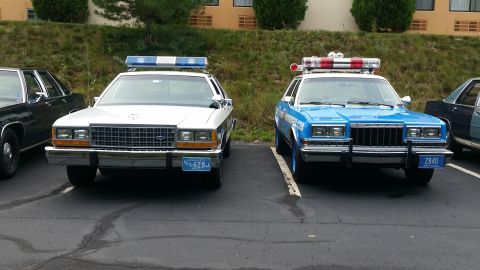 1991 Ford LTD Crown Victoria - A white 1991 Ford Crown Victoria and a blue police car are parked side-by-side in a lot.