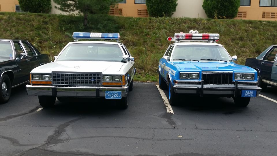 Two police cars parked in a lot, one white and one blue. Both have light bars on top