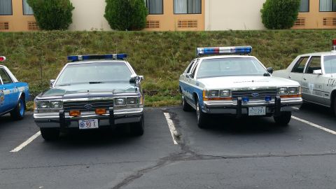 1991 Ford LTD Crown Victoria - Two police cars, a dark green Ford Crown Victoria and a white Ford Crown Victoria, are parked side-by-side.