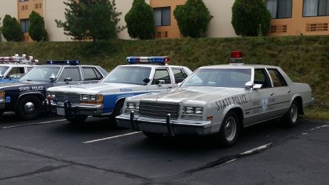1991 Ford LTD Crown Victoria - Multiple police vehicles are lined up in a parking lot, some with light bars on top.