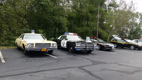 1991 Ford LTD Crown Victoria - Several police cars are parked in a lot, including a vintage cream-colored model and newer black models.