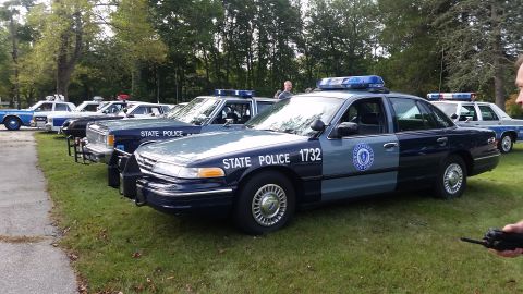 1991 Ford LTD Crown Victoria - Multiple State Police vehicles lined up on a grassy area with trees in the background.