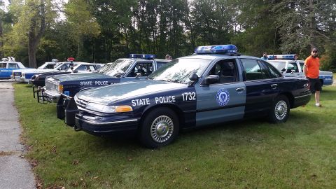 1991 Ford LTD Crown Victoria - A line of state police vehicles are parked on a grassy area with trees in the background.