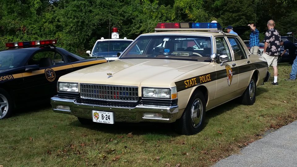 Tan state police car with light bar, parked next to a black state trooper car