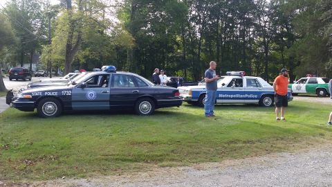 1991 Ford LTD Crown Victoria - Several police cars, including a "STATE POLICE" vehicle, are parked on a grassy area with trees in the background.