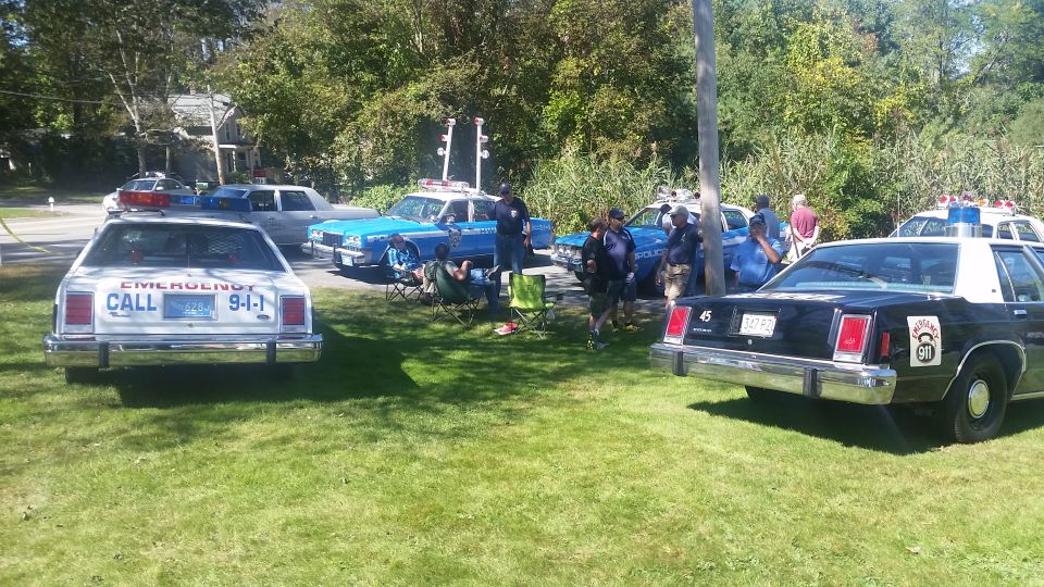 1991 Ford Crown Victoria police car rear view, white with 'EMERGENCY CALL 9-1-1' on trunk. Other police cars visible