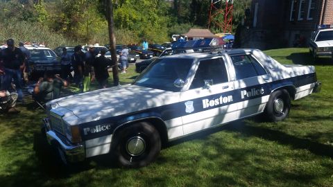 1991 Ford LTD Crown Victoria - A Boston Police car is seen parked on a grassy area with other vehicles and people in the background.