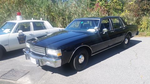 1991 Ford LTD Crown Victoria - A black sedan is parked next to a white state police car. The sedan appears to be from the 1980s.