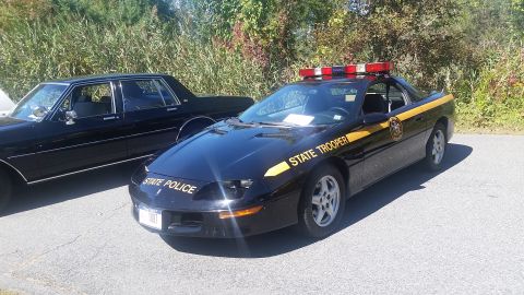 1991 Ford LTD Crown Victoria - A black Chevrolet Camaro is marked as a state trooper vehicle, parked next to a dark sedan.