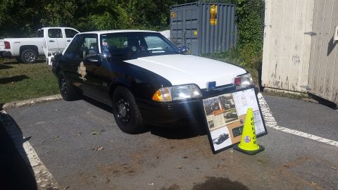 1991 Ford LTD Crown Victoria - Police car on display with informational board and traffic cone. A pickup truck is parked nearby.