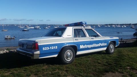 1991 Ford LTD Crown Victoria - A police car with 'Metropolitan Police' text and a phone number is parked on a grassy area near a body of water filled with sailboats.