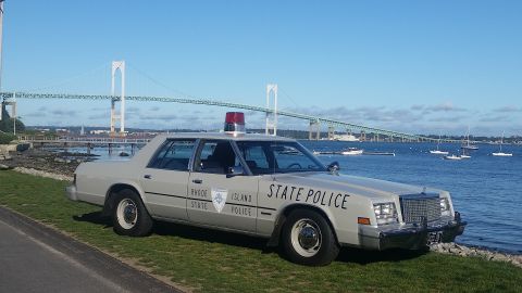 1991 Ford LTD Crown Victoria - A vintage Rhode Island State Police car sits parked on the grass near the water, with a large bridge spanning the bay in the distance.