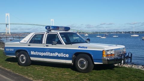 1991 Ford LTD Crown Victoria - A 1991 Ford Crown Victoria police car, marked 'Metropolitan Police', is parked on a grassy area next to the water with a bridge and sailboa…