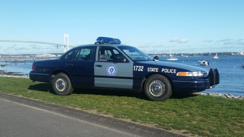1991 Ford LTD Crown Victoria - A Massachusetts State Police car is parked on the grass near the water, with boats and a bridge visible.
