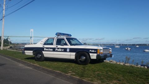 1991 Ford LTD Crown Victoria - A Boston Police car is parked on a grassy area next to the water, with boats visible in the distance.