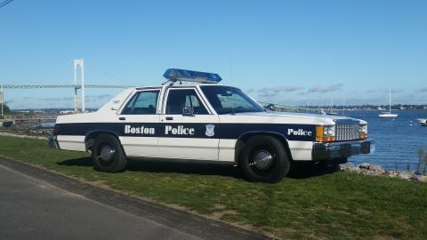 1991 Ford LTD Crown Victoria - A Boston Police car is parked by the water with a bridge visible in the distance.