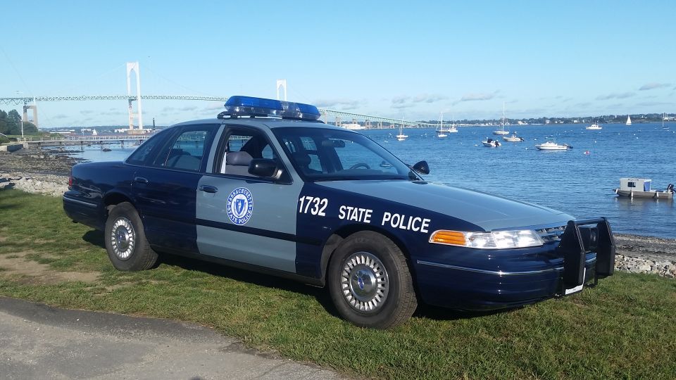 Dark blue and light blue police car with "STATE POLICE" text and number 1732, parked on grass by the water with a bridge in the background