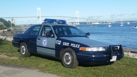 1991 Ford LTD Crown Victoria - A 1991 Ford Crown Victoria police car, marked '1732 STATE POLICE', sits parked on a grassy area next to a body of water with a bridge in th…