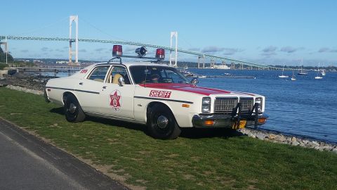 1991 Ford LTD Crown Victoria - A vintage sheriff car is parked on a grassy area next to the water, with a large bridge spanning the bay in the distance.