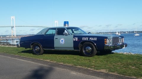 1991 Ford LTD Crown Victoria - A vintage Massachusetts State Police car is parked on a grassy area near the water, with a large bridge visible in the distance.
