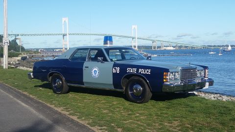 1991 Ford LTD Crown Victoria - A vintage Massachusetts State Police car is parked on a grassy area next to the water, with a large bridge spanning the bay in the distance.