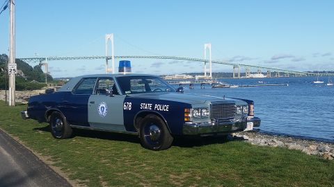 1991 Ford LTD Crown Victoria - A two-tone blue and grey police car is parked on a grassy area next to the water, with a large bridge visible in the distance.