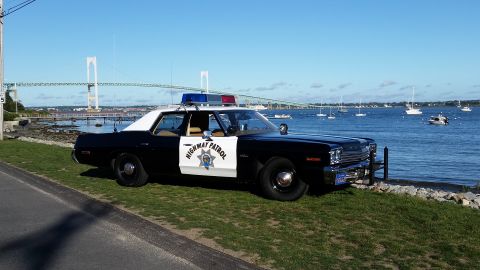 1991 Ford LTD Crown Victoria - A vintage highway patrol car is parked near the water with sailboats in the distance and a large bridge overhead.