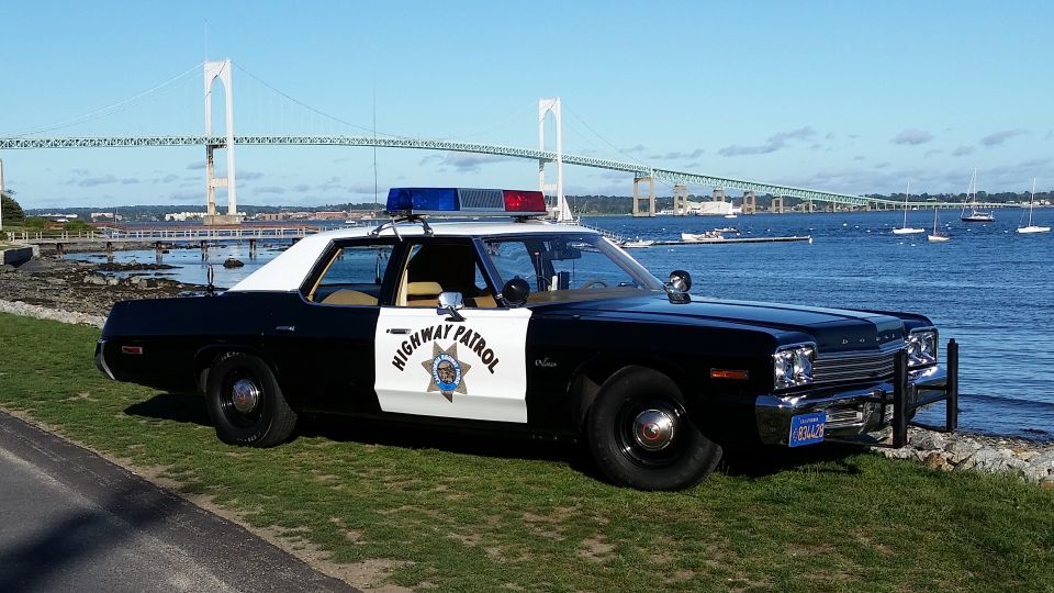 Black and white police car with "HIGHWAY PATROL" on the door, parked on grass near the water. A large bridge is visible in the background