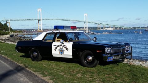 1991 Ford LTD Crown Victoria - A vintage black and white police car sits on a grassy area near the water, with a large bridge visible in the distance.