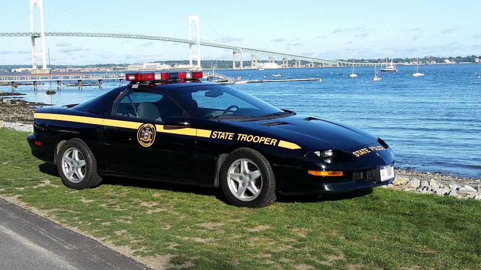 Black police car with yellow stripe and "STATE TROOPER" text parked by the water with a bridge in the background