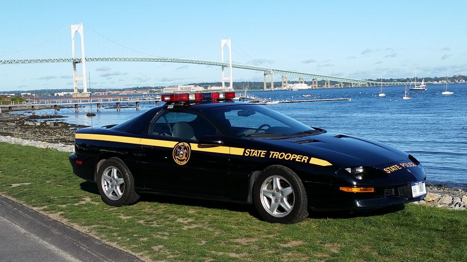 Black state trooper car with yellow stripe and "STATE TROOPER" text parked by the water with a bridge in the background