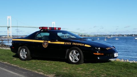 1991 Ford LTD Crown Victoria - A black police car with a yellow stripe and "STATE TROOPER" text is parked by the water with a bridge in the distance.