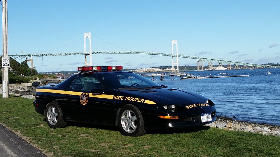 Black police car with yellow stripe and "STATE TROOPER" text, parked on grass near water with a bridge in the background