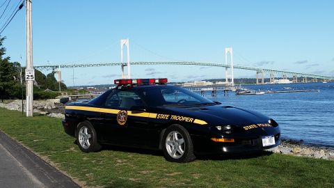 1991 Ford LTD Crown Victoria - A black police car with a yellow stripe and "STATE TROOPER" text is parked on the grass near the water, with a large bridge visible in the…