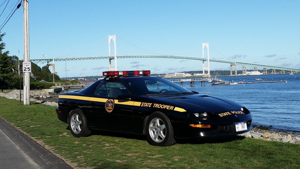 Black police car with yellow stripe and "STATE TROOPER" text parked by the water with a bridge in the background